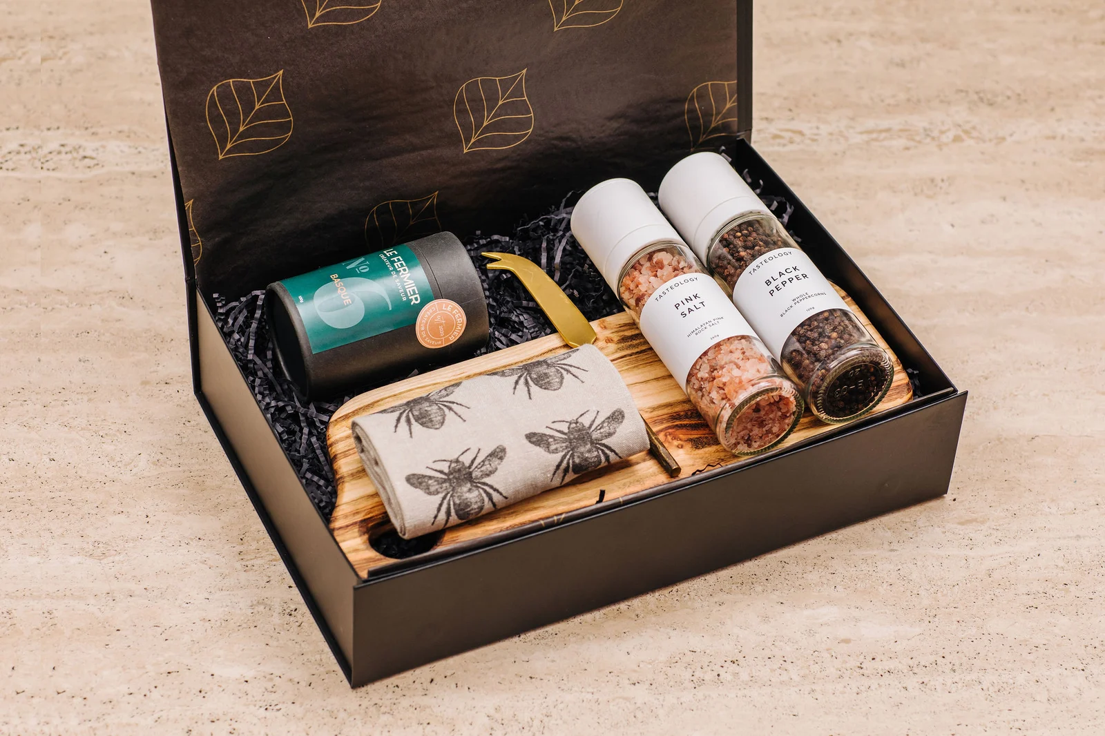 A gourmet gift hamper with a wooden serving board, bee-print cloth and salt and pepper grinders